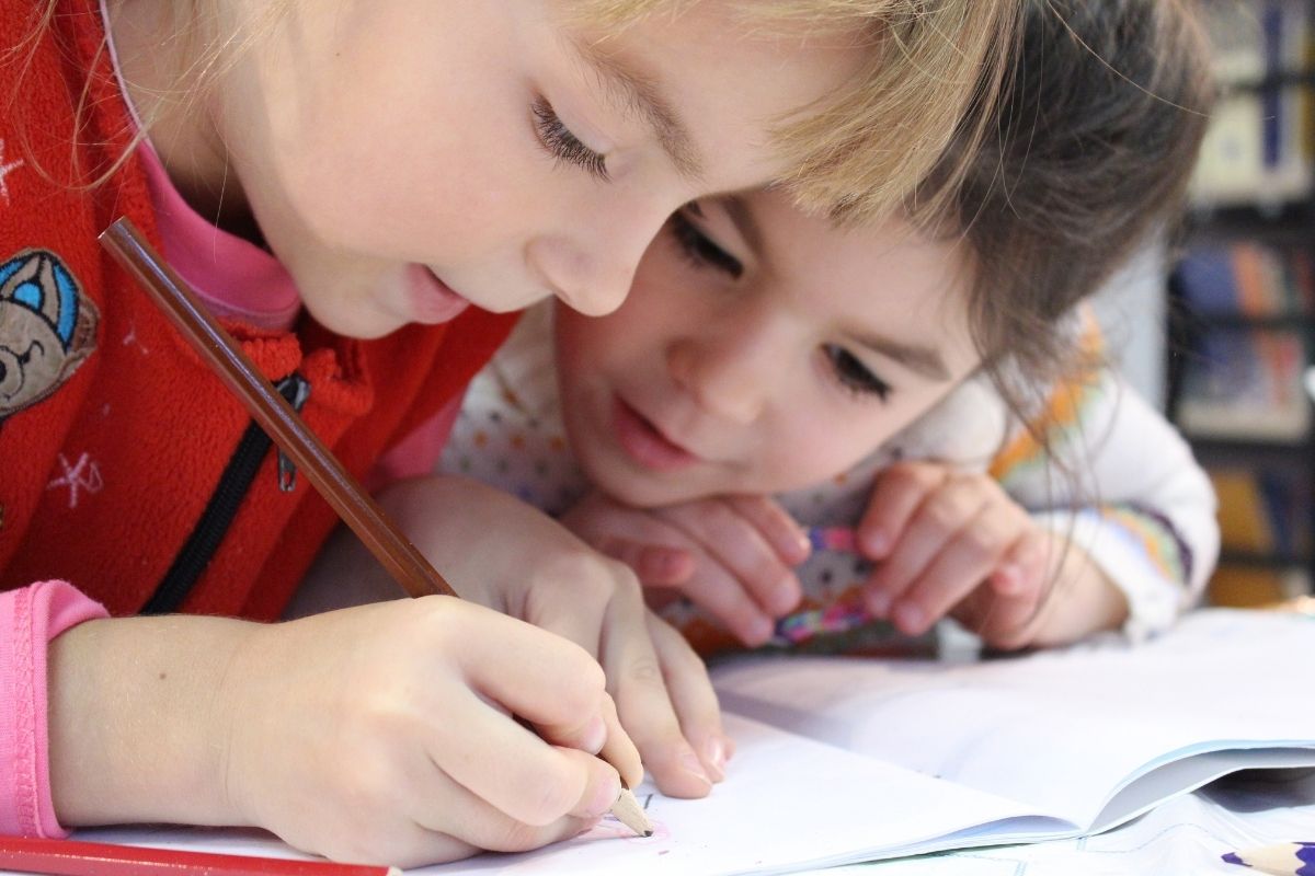 Two girls, one blonde, the other brunette, working together on their workbook in a classroom of an infant school.